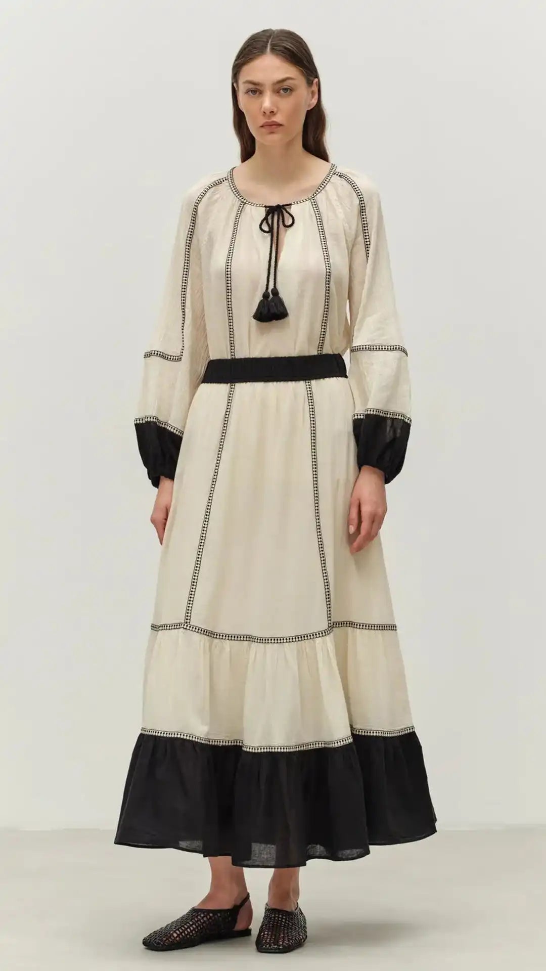 An off-white tiered maxi dress with black trim, long sleeves, and a tassel tie-neck.