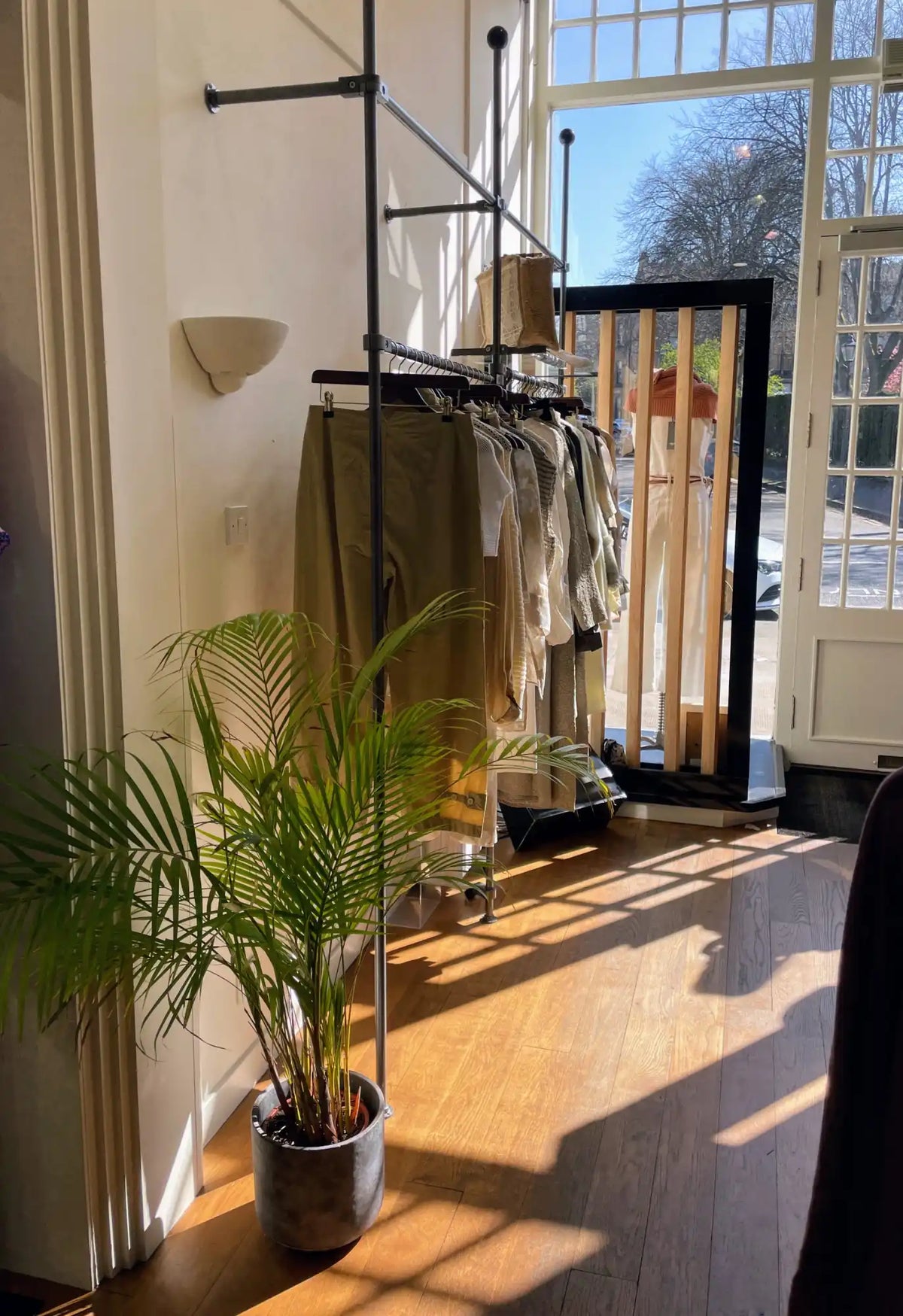 A tall, dark metal clothing rack displaying various neutral-toned garments, including pants and shirts.